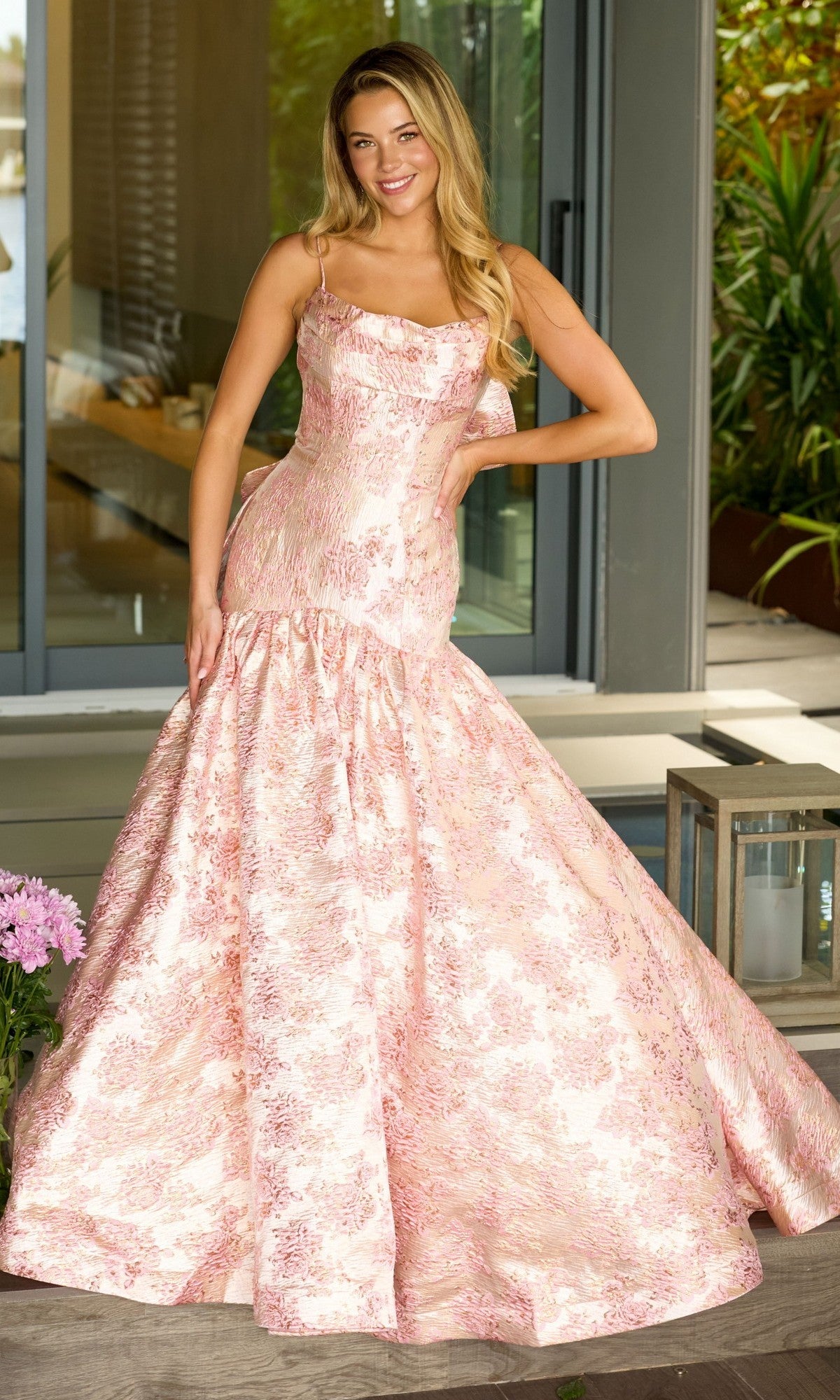 A woman stands on a porch in the Ava Presley 26419 Drop-Waist Long Print Prom Gown with thin straps, confidently posing with one hand on her hip.